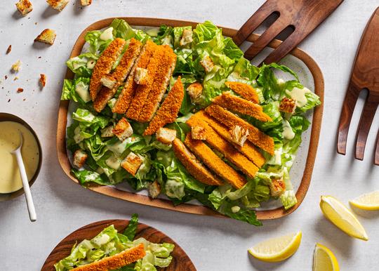 A photo of a salad, with breaded chicken on top, surrounded by lemon wedges on a clean white table.